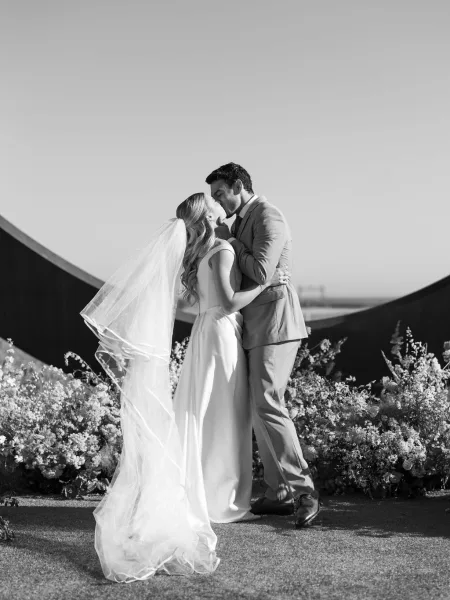 Wedding kiss portrait of bride and groom kissing in a close embrace, her long veil blowing over a strapless gown on an outdoor lawn by flowers