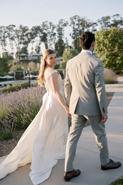 Couple portrait of bride and groom holding hands walking down a garden pathway, bride looking back as her train trails by lavender at golden hour