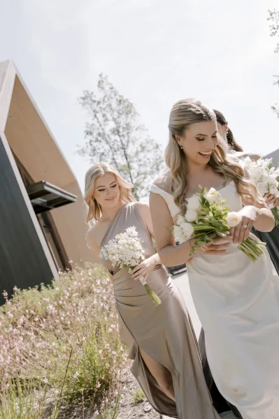 Bride portrait with a white rose bridal bouquet, laughing as a bridesmaid walks behind on an outdoor walkway by modern architecture and wildflowers