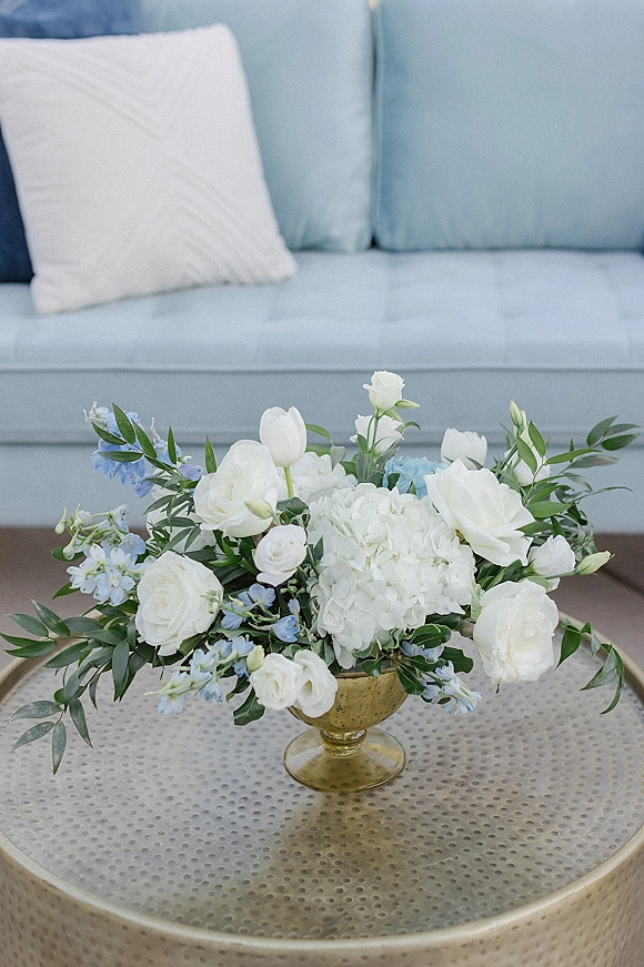Wedding centerpiece with white roses and hydrangea rose centerpiece in a gold bowl vase, accented with blue flowers and greenery on a round metal coffee table