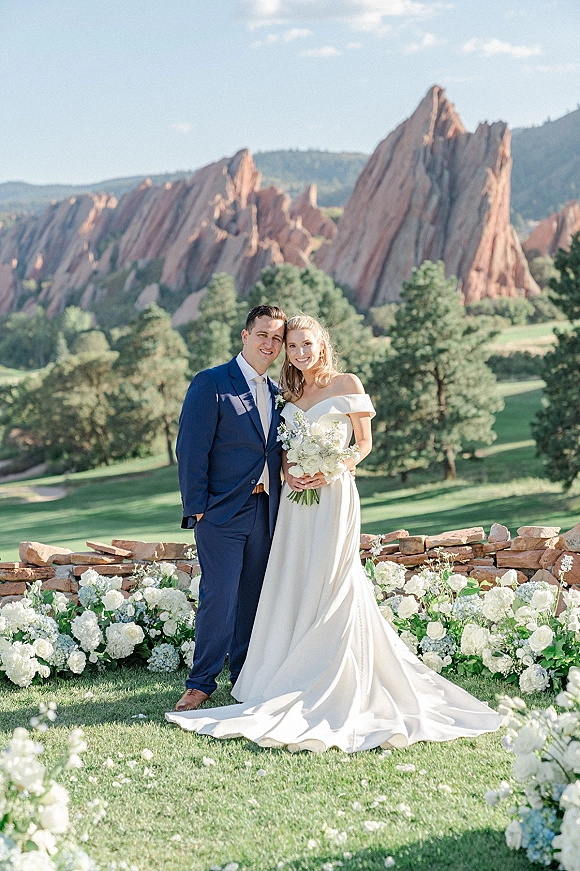 Couple portrait of bride in off the shoulder dress holding a white floral bouquet beside groom in a blue suit by mountain rocks and trees
