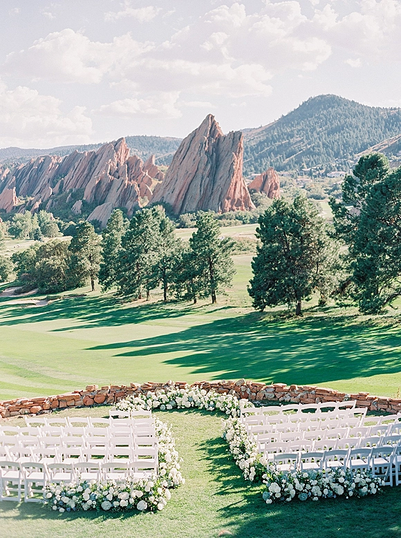Outdoor ceremony setup with mountain wedding ceremony seating, white folding chairs in a curve along a flower-lined aisle beside a stone wall
