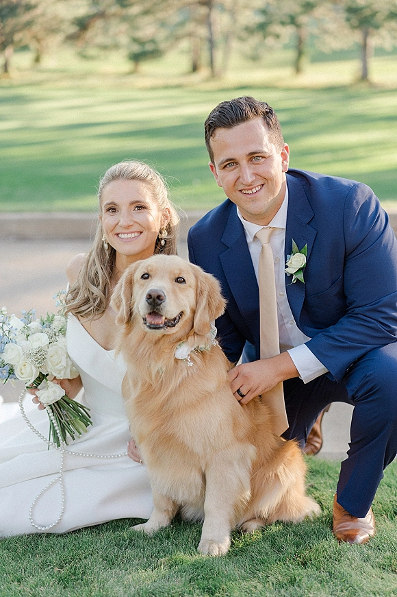 Couple portrait of bride and groom with dog, bride holding a white rose bouquet as they kneel on a park lawn by trees and path