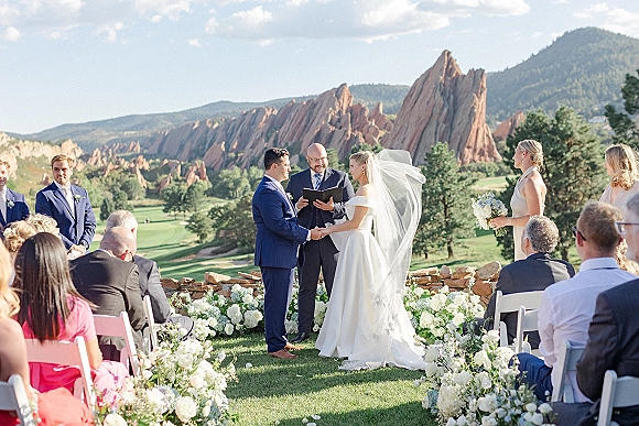 Wedding ceremony with bride and groom holding hands as officiant reads, bridal veil flowing before red rock mountains and stone wall backdrop