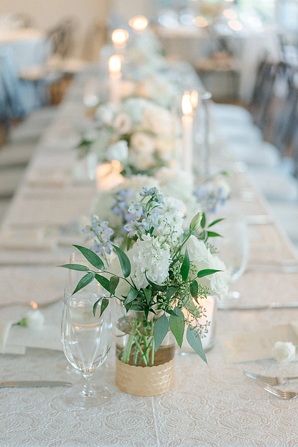Reception tablescape with wedding table centerpiece of white flowers and greenery, taper candles and glass goblets on patterned linens in a softly lit room