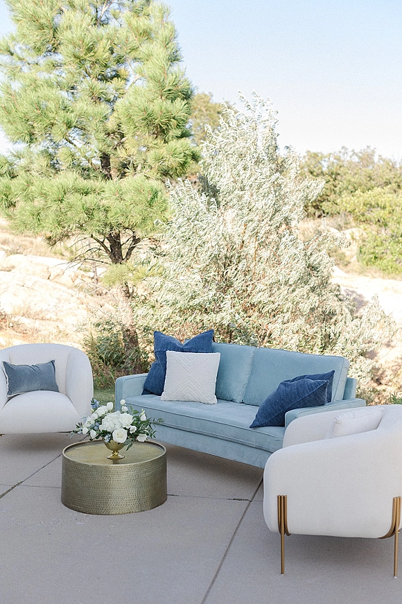 Wedding lounge seating with a blue sofa and white armchairs around a round brass coffee table with floral accents on an outdoor patio