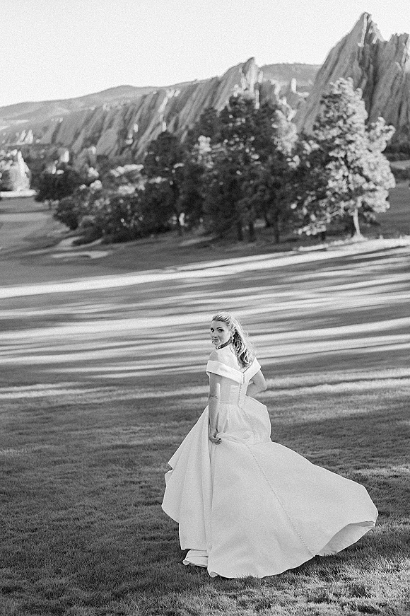 Bridal portrait in a grassy field, bride looking back in an off-the-shoulder gown with long train, set against mountain rocks and trees