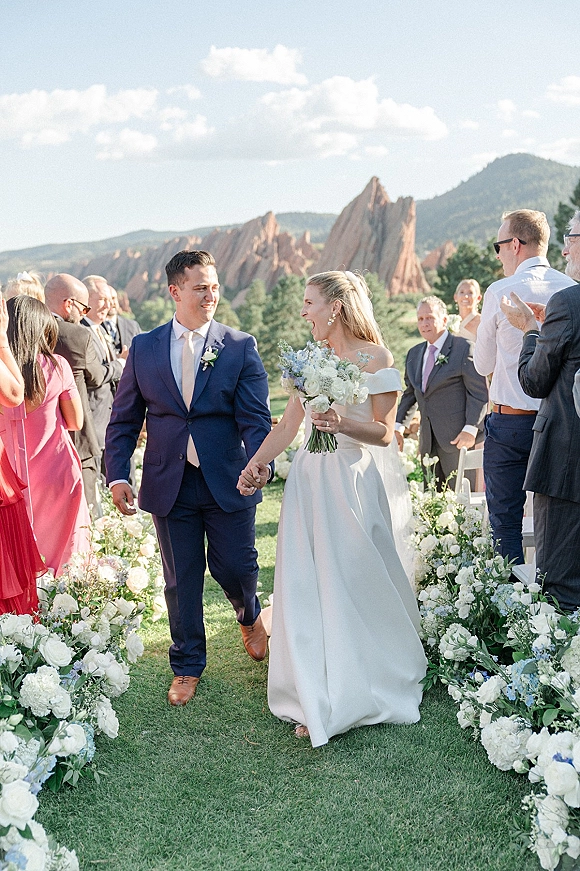 Wedding recessional as bride and groom walk hand in hand down the aisle, bride laughing with bouquet, mountains and rocks behind them