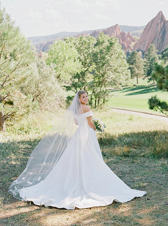 Bridal portrait of a bride in an off the shoulder wedding dress with cathedral veil and white bouquet, standing in a meadow with mountains behind