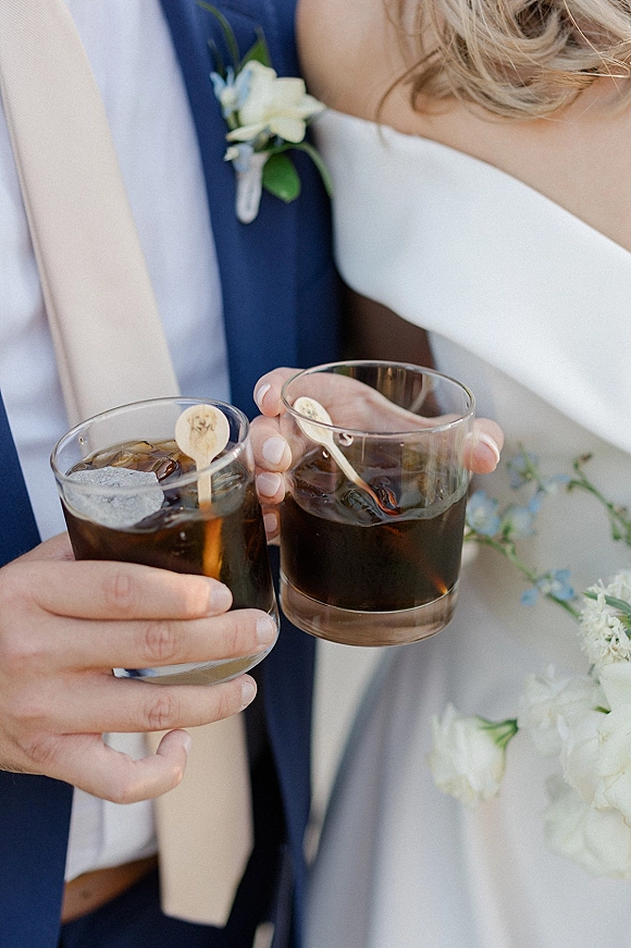 Wedding drink toast as bride and groom clink iced dark cocktails in rocks glasses, bouquet and boutonniere visible against blurred florals