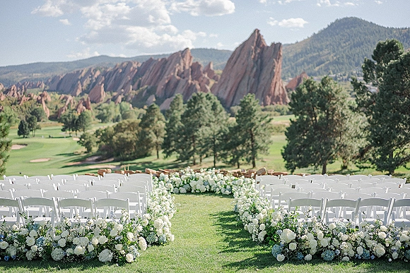Outdoor ceremony setup with wedding aisle flowers lining a grassy aisle, white folding chairs, and hydrangeas beneath red rock mountains