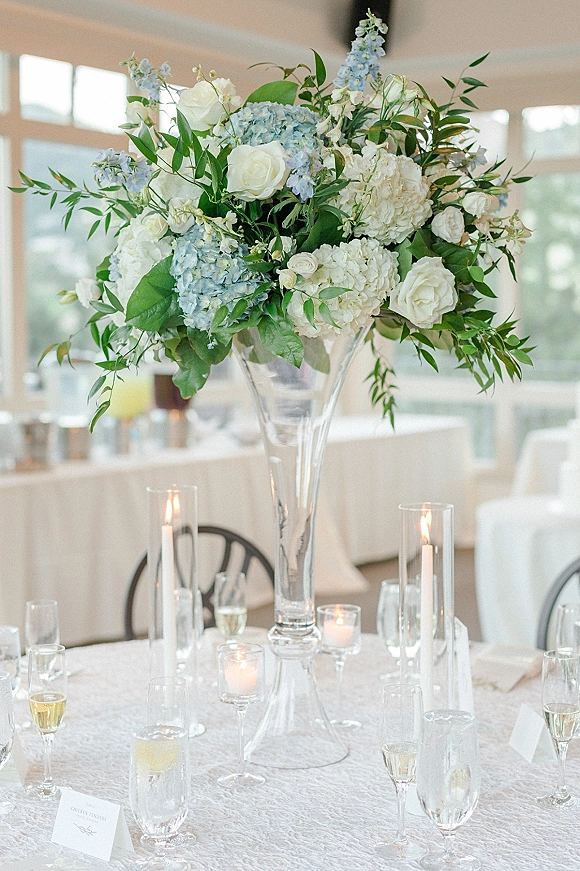 Reception centerpiece with a tall floral centerpiece of white roses and blue hydrangea in a glass vase, accented by taper candles on a linen table
