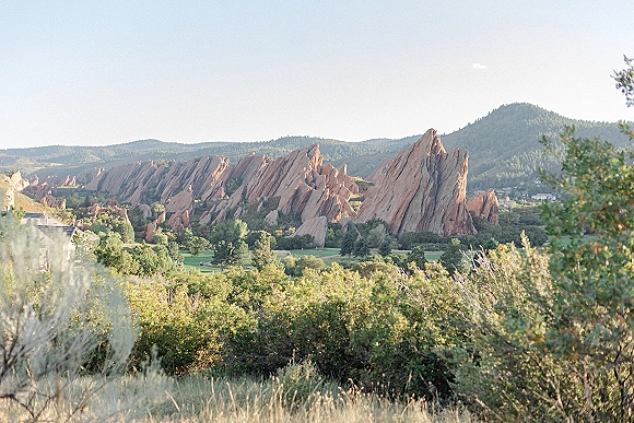 Mountain landscape with a red rock ridge above evergreen forest and foothills, scrubby grass in the foreground under clear sky