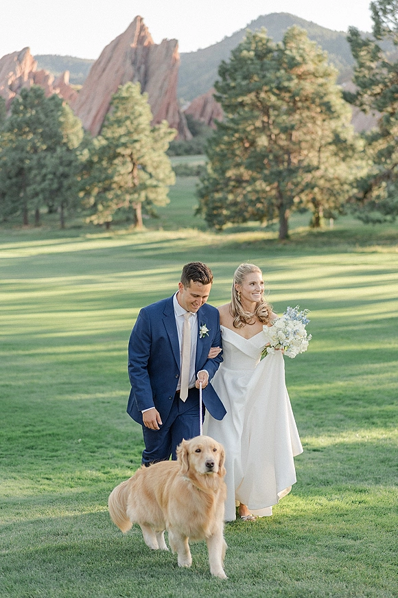 Couple portrait of bride and groom with dog, walking arm in arm on a mountain lawn, bride holding bouquet and leash
