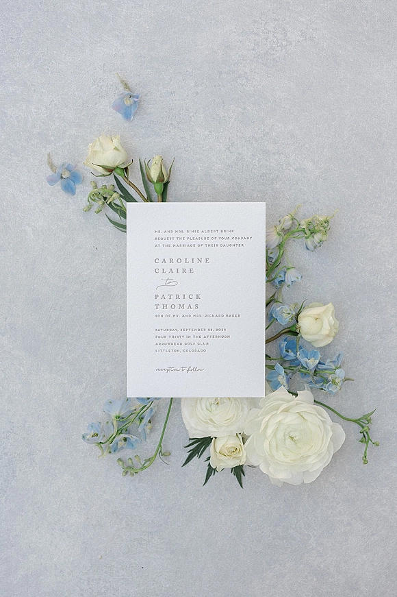 Wedding invitation flat lay with white roses, blue flowers, and greenery arranged around an invitation card on a gray textured surface