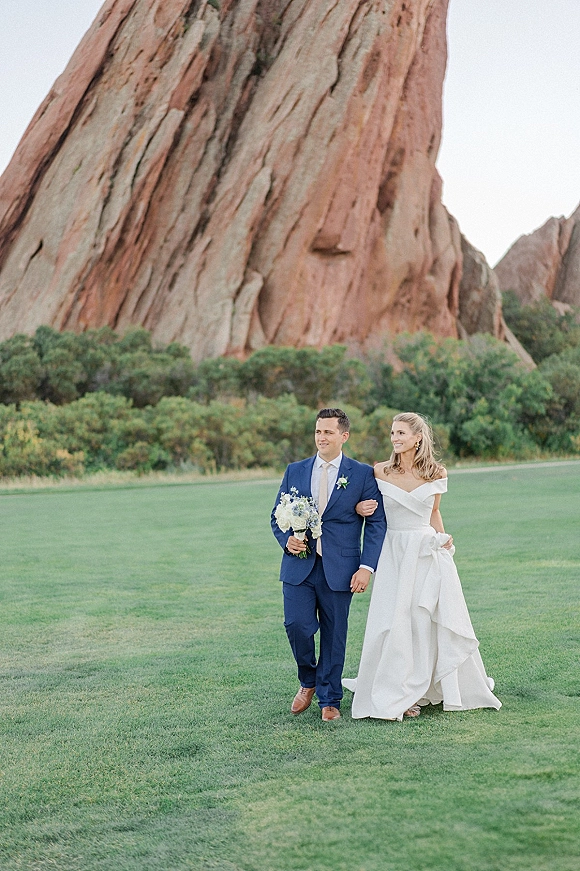 Couple portrait of bride and groom walking arm in arm on a green lawn, her off-shoulder gown and bouquet, his blue suit, red rock cliffs behind