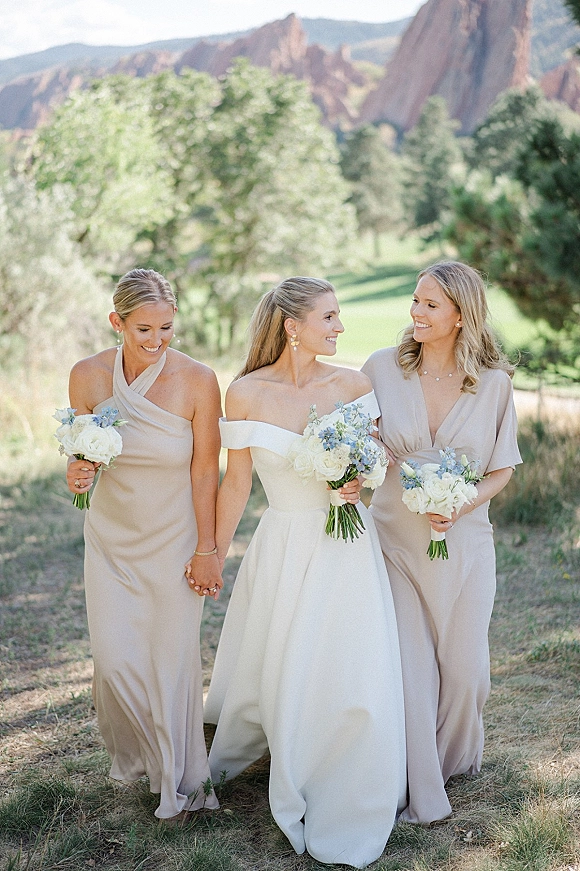 Bride with bridesmaids walking with bride hand in hand, holding white and blue bouquets against mountain cliffs and trees outdoors
