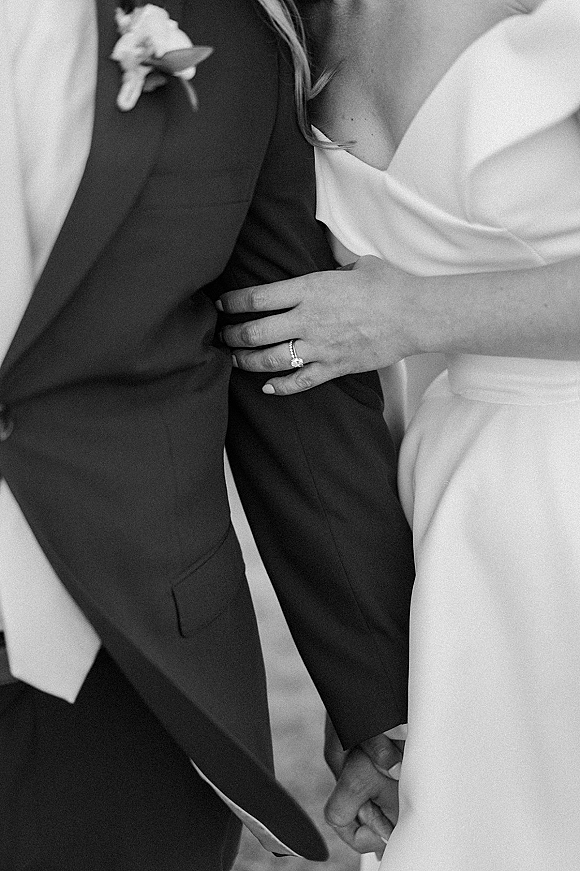 Couple portrait with bride holding groom’s arm, wedding ring close up showing diamond solitaire and band stack against a neutral backdrop