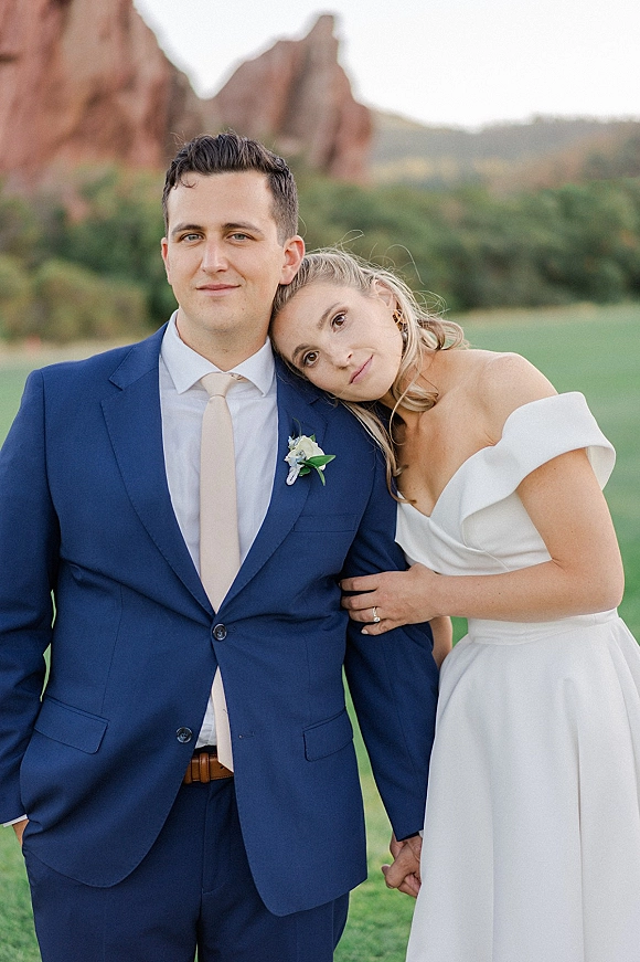 Couple portrait of bride leaning on groom, holding hands, her off-the-shoulder dress beside his blue suit with red rock formations behind