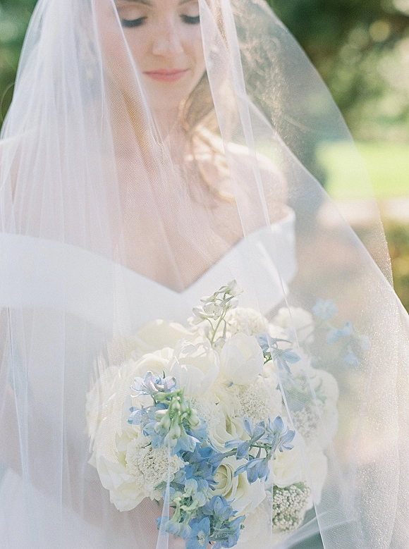 Bridal portrait of a bride with veil over face holding a white and blue bouquet, looking down in sunlit outdoor greenery bokeh