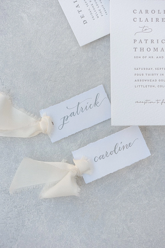 Wedding place cards with torn-edge name tags and silk ribbon bows beside an invitation on light gray linen tablecloth flat lay