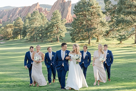 Wedding party portrait with bride and groom walking with bridesmaids holding white bouquets and groomsmen in navy suits, mountains behind