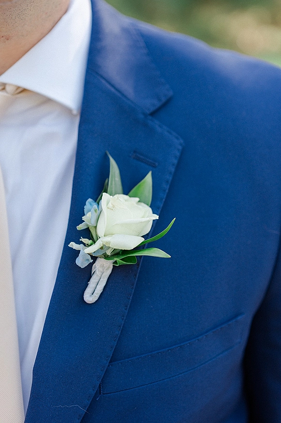 Wedding boutonniere with a white rose boutonniere, greenery, and blue accent flowers pinned to a blue suit jacket against outdoor bokeh greenery