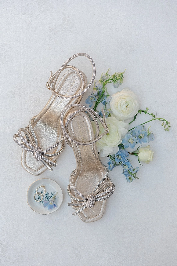 Bridal shoes in a wedding shoes flat lay with metallic strappy heels, pearl-beaded ankle straps, bow detail, and blue flowers on white texture