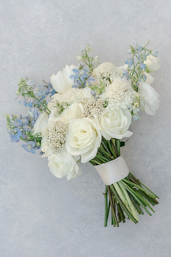 Bridal bouquet with white and blue bouquet tones, featuring roses, ranunculus, delphinium, and ribbon wrap against a gray backdrop