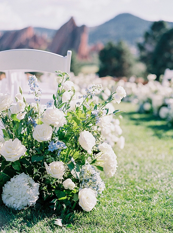 Ceremony aisle decor with white folding chairs and white rose, hydrangea ground florals along a grass lawn, mountains and rocks beyond