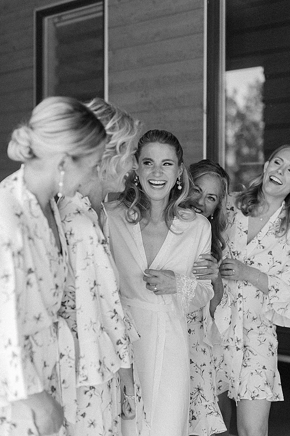Getting ready moment as bride with bridesmaids laughs in bridal robes with lace cuffs by a window against a wood wall backdrop