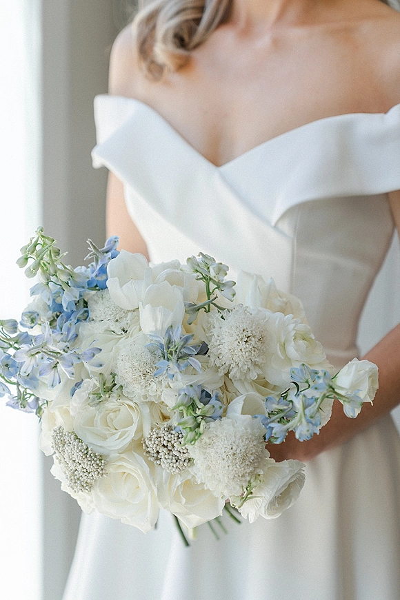 Bridal bouquet of white roses with blue delphinium accents and baby's breath, held against an off-the-shoulder dress in window light