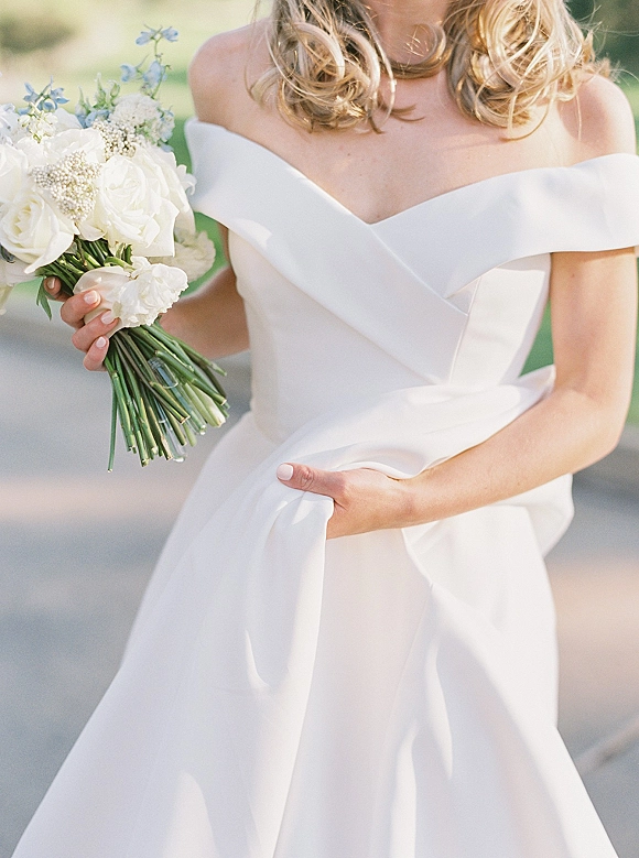 Wedding dress close-up with off the shoulder wedding dress wrap bodice as bride holds white rose bouquet with baby’s breath and blue flowers on a walkway