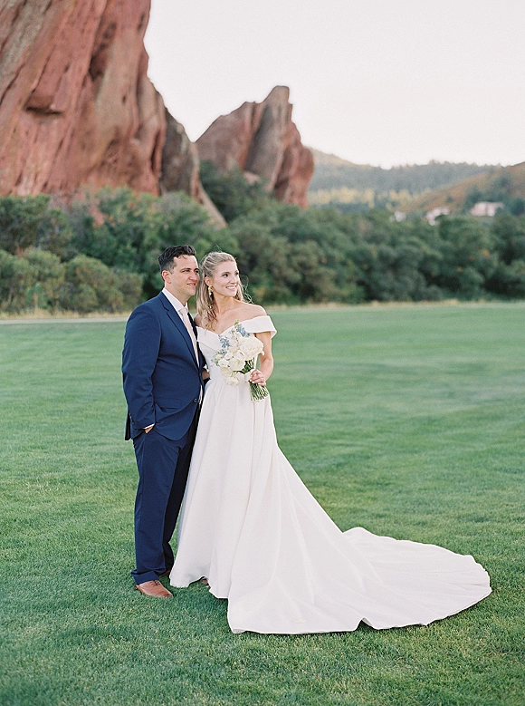 Couple portrait of bride and groom standing pose on a green lawn, bride in off-shoulder gown holding bouquet beside groom in blue suit near rocky hills
