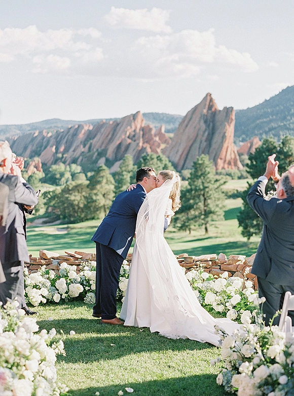 Wedding kiss as bride in flowing veil and dress embraces groom in suit, framed by aisle florals and mountain stone wall backdrop
