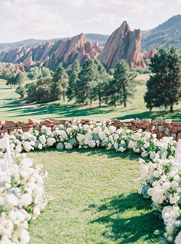 Outdoor ceremony setup with wedding ceremony aisle flowers and white chair rows, accented by lush ground florals with mountain backdrop