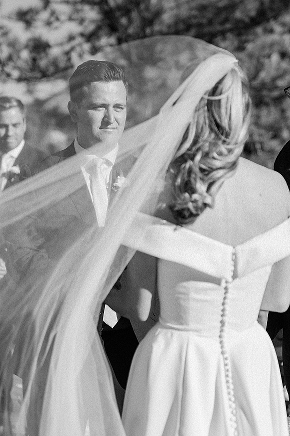 Wedding ceremony moment as bride walks to groom, veil blowing behind her, while he waits in suit with boutonniere among guests and trees