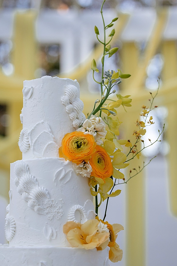 Wedding cake with floral wedding cake topper of yellow and orange blooms on textured white buttercream tiers against soft drapery bokeh