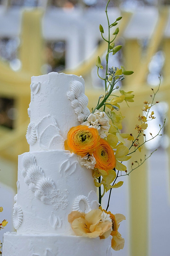 Wedding cake with white buttercream frosting and piped icing, topped with orange ranunculus and yellow orchids against soft bokeh lights