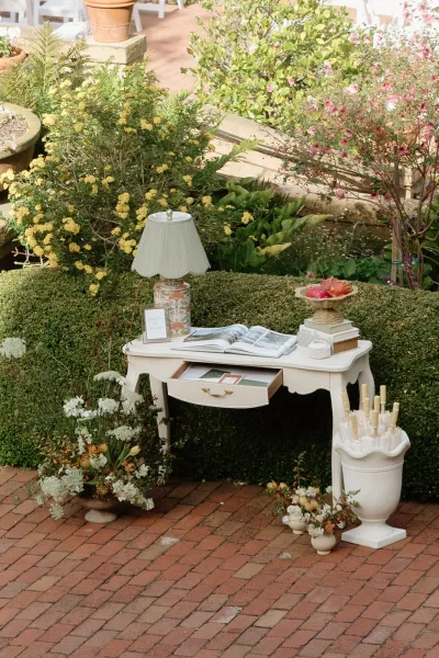 wedding guest book table featuring welcome table decor and florals on a brick patio beside garden hedges and white chairs