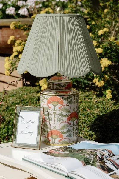 Wedding table lamp serving as a lamp centerpiece, its pleated lampshade set against garden greenery with yellow flowers and a terracotta planter.