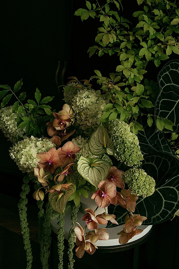 Wedding centerpiece with green hydrangeas, orchids, and anthurium spilling from a ceramic vase, trailing amaranthus against a dark leafy backdrop