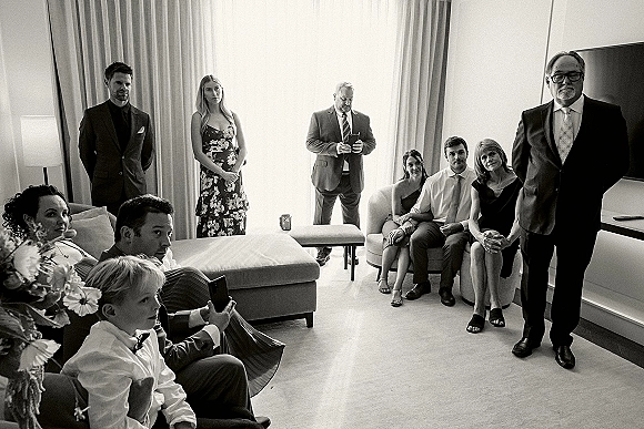 Wedding guests in a living room, some seated on the sofa and others standing, wearing suits and dresses while one holds a smartphone