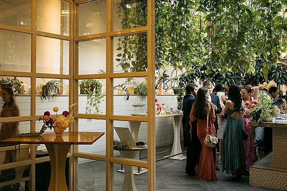 Wedding cocktail hour with wedding guests mingling around highboy tables, candlelit floral centerpieces, and greenery in a glass-partitioned patio