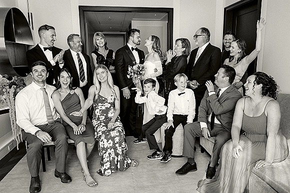 Wedding group photo of bride and groom with family, bouquet accent, laughing indoors by a doorway with mirror, chairs, and floral decor