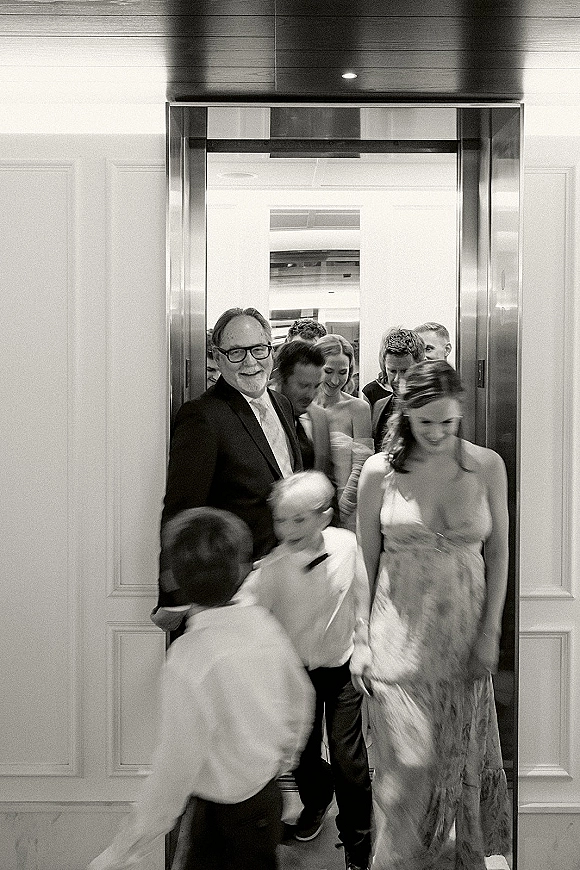 Wedding guests in formalwear exiting an elevator, smiling in a hotel hallway under ceiling lights, with suits, eyeglasses, and a boutonniere