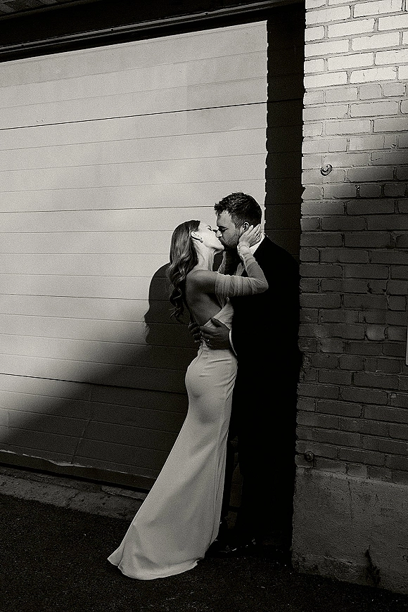 Wedding kiss portrait of bride and groom kissing in a close embrace, her long-sleeve low-back gown and his boutonniere in harsh alley shadows