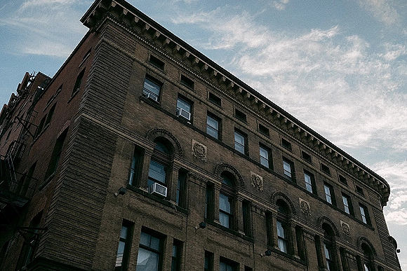 Historic brick building with arched windows and stone detailing, viewed from a corner upward angle with a fire escape against a cloudy sky