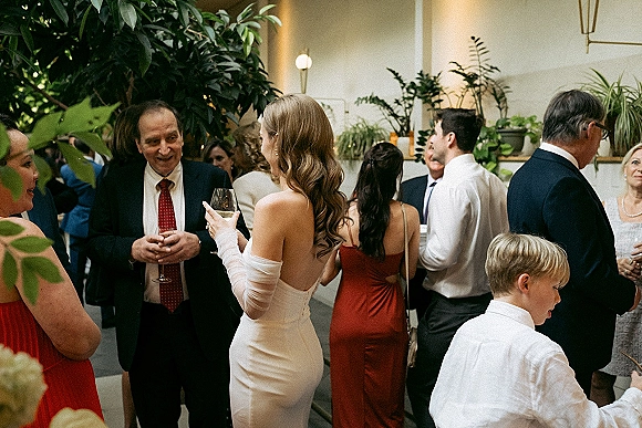 Wedding cocktail hour as bride in sheer opera gloves holds a wine glass, chatting with guests in a lush indoor greenhouse with potted plants