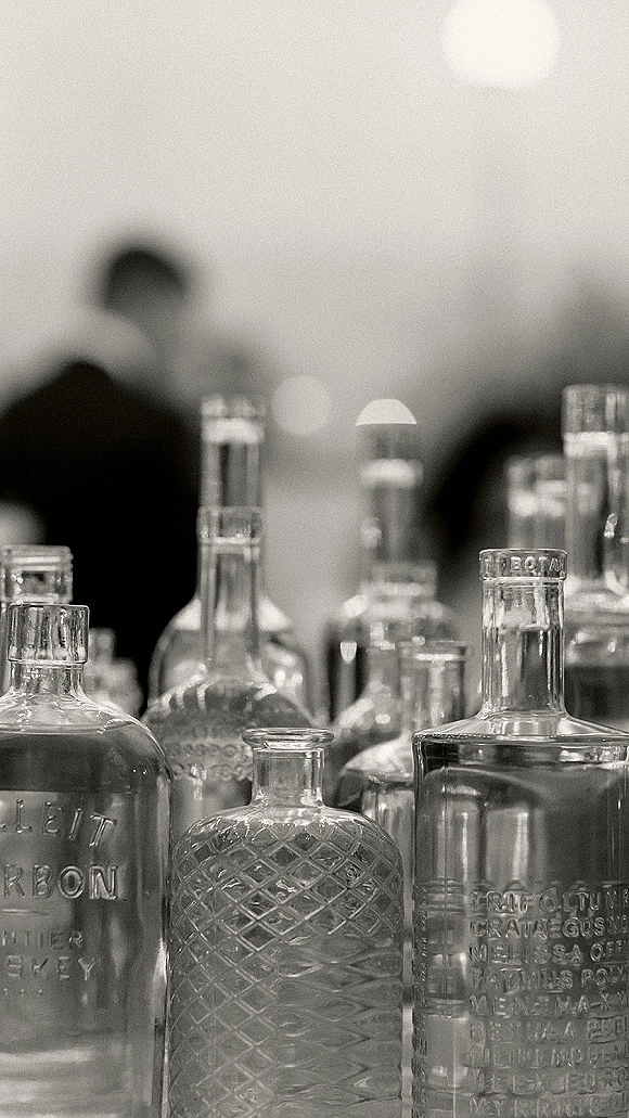 Wedding bar setup with glass liquor bottles, decanters, and bar glassware on a reception counter, with blurred guests in warm indoor lighting